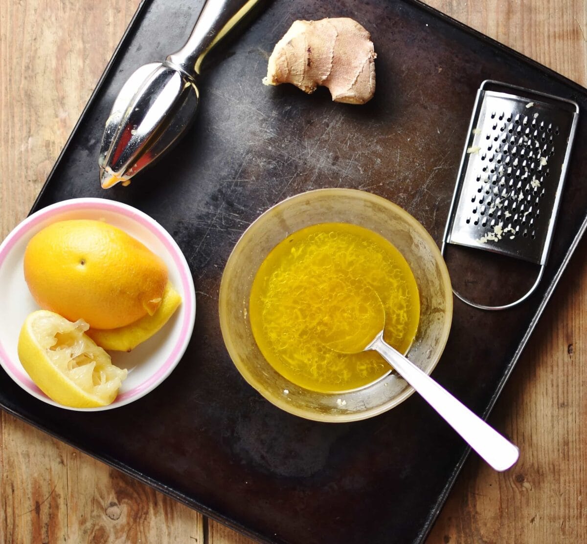 Salad dressing with spoon in bowl, zester, ginger, juicer and orange on top of small white plate.