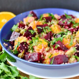 Side view of beets quinoa salad in blue bowl on white plate, with herbs and oranges in background.