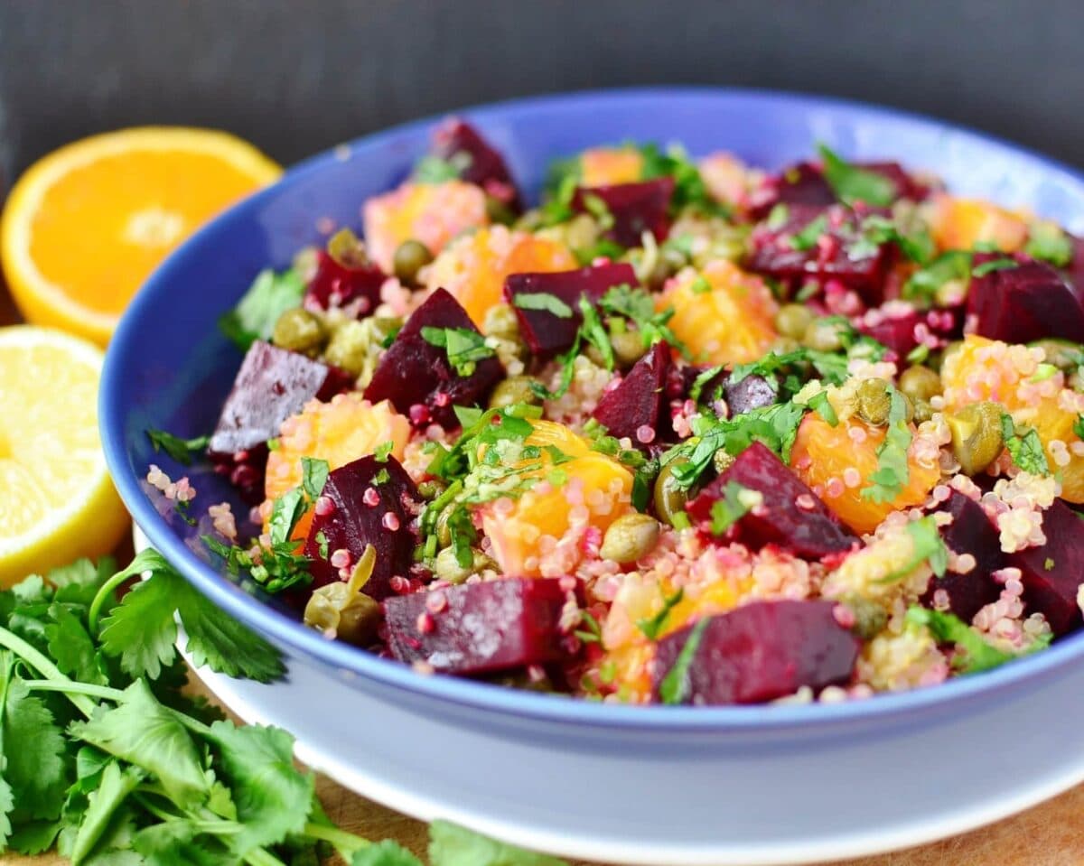 Side view of beets salad with quinoa in blue bowl on white plate, with herbs and oranges in background.