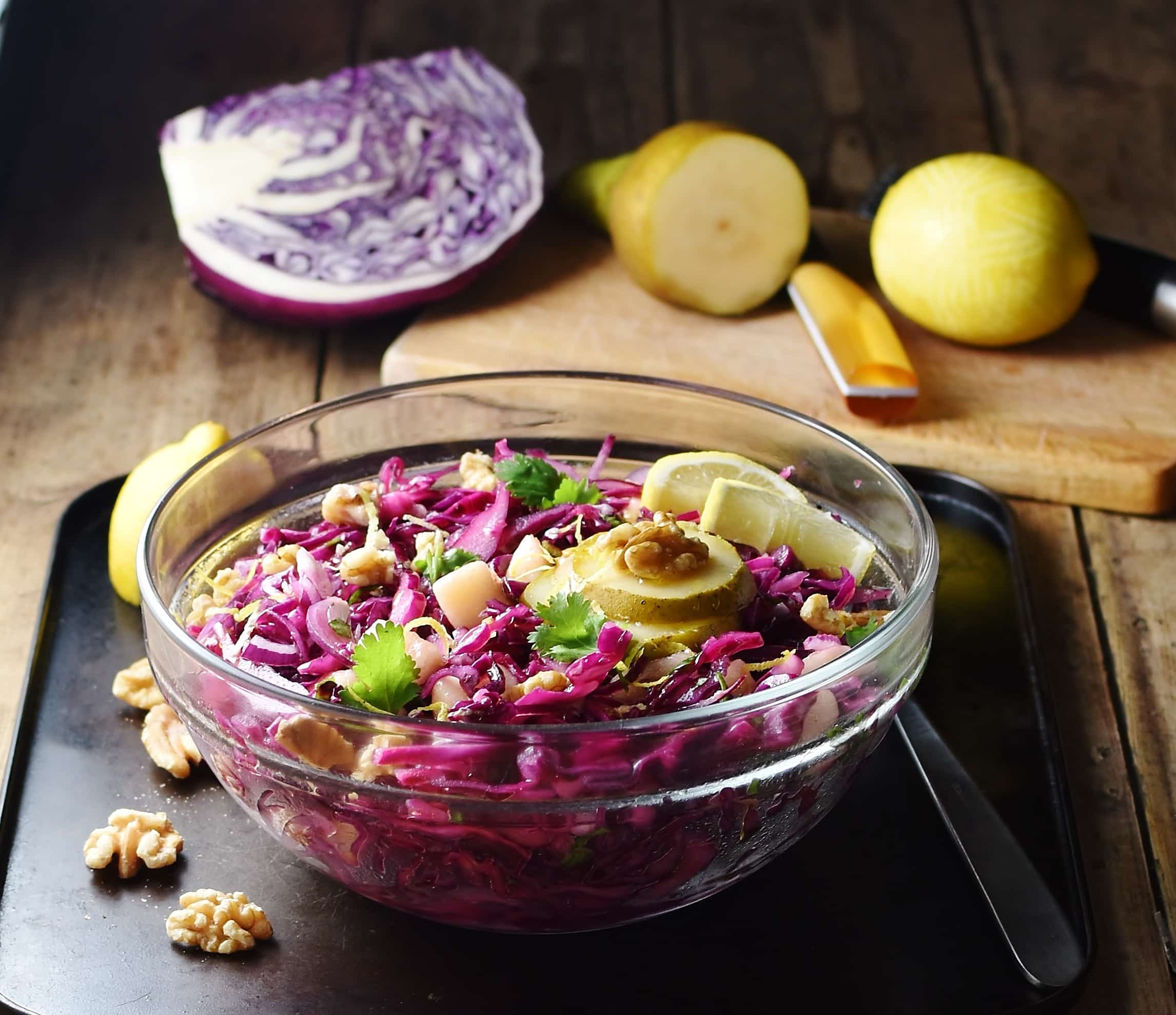 Shredded red cabbage with pear, walnuts and herbs in mixing bowl with spoon to the right, lemon ,pear, red cabbage piece and knife in background.