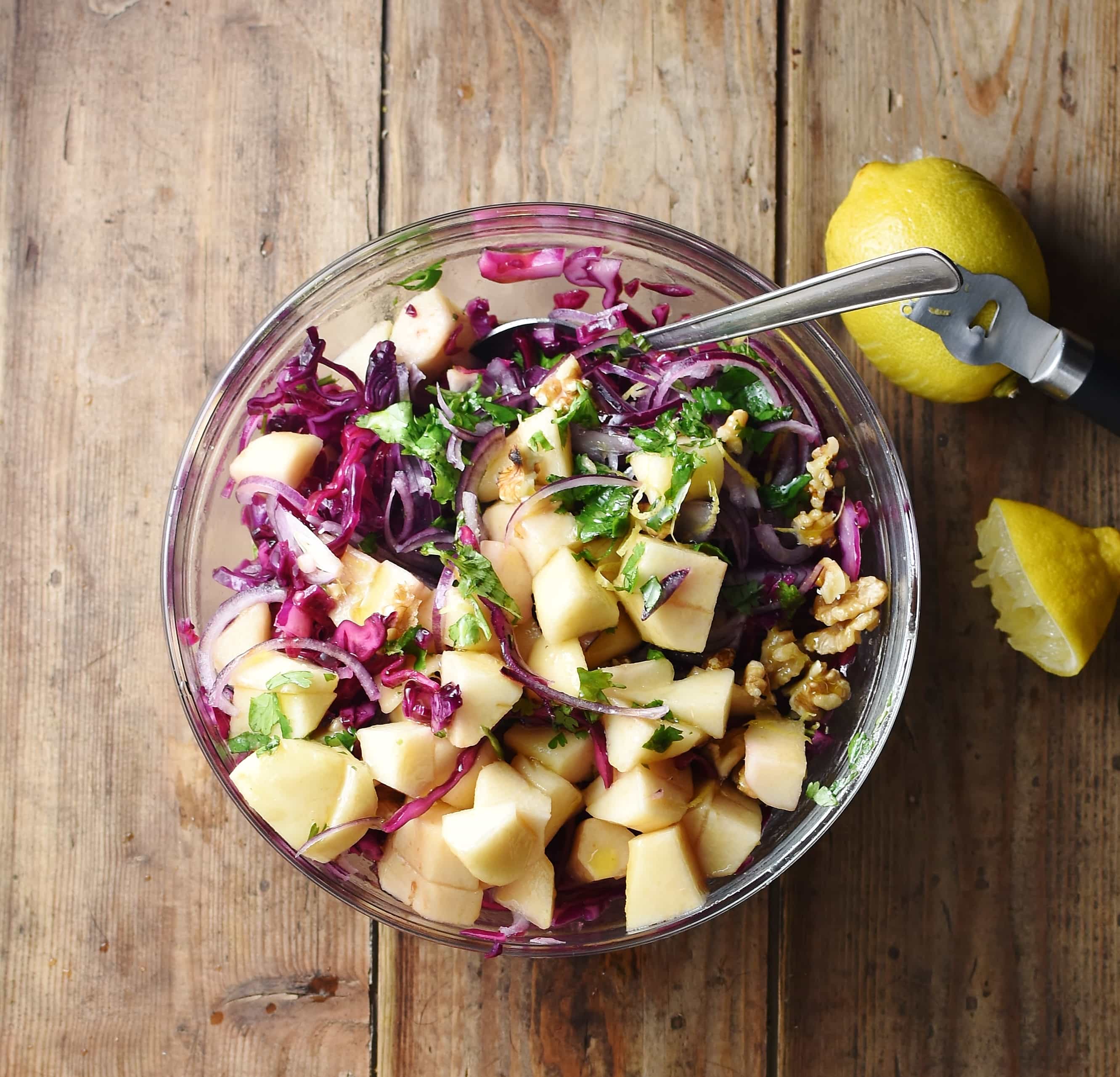 Chopped pear, walnuts, herbs and red cabbage in mixing bowl with spoon and lemons to the right.