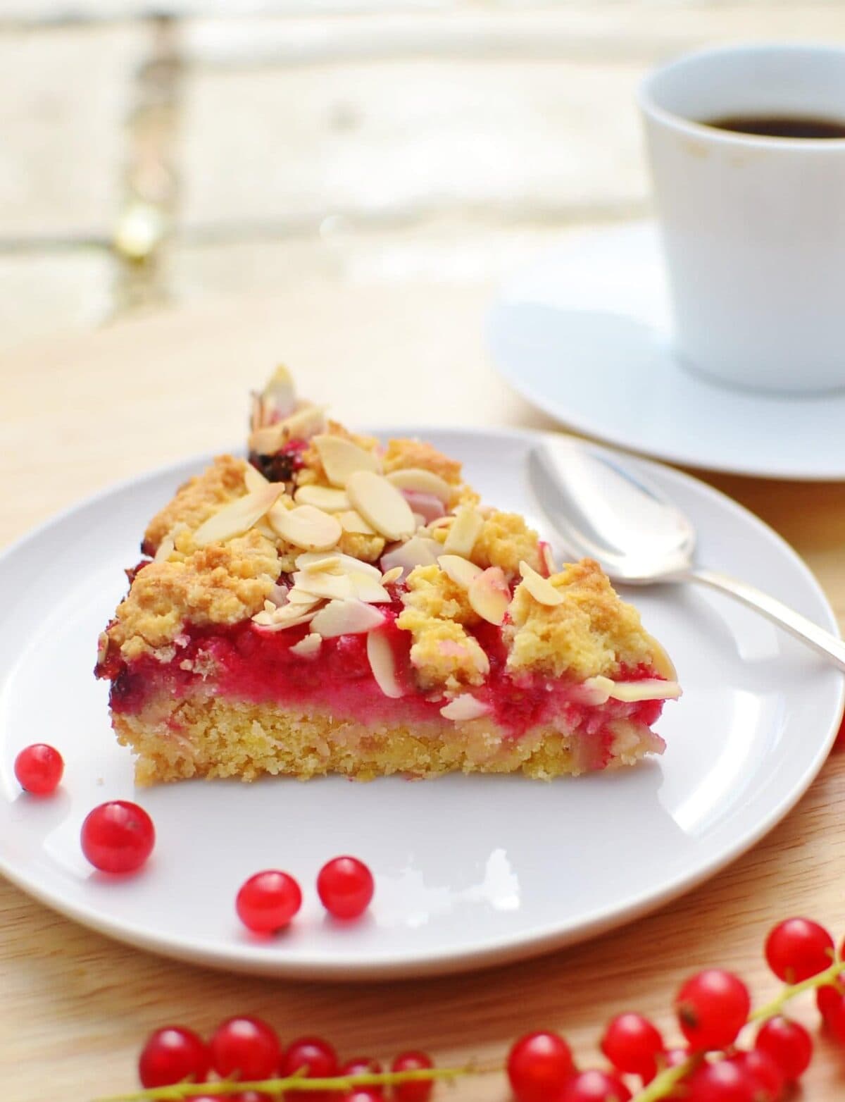 Side view of almond redcurrant cake slice on white plate with redcurrants on wooden surface with white cup and saucer in background.