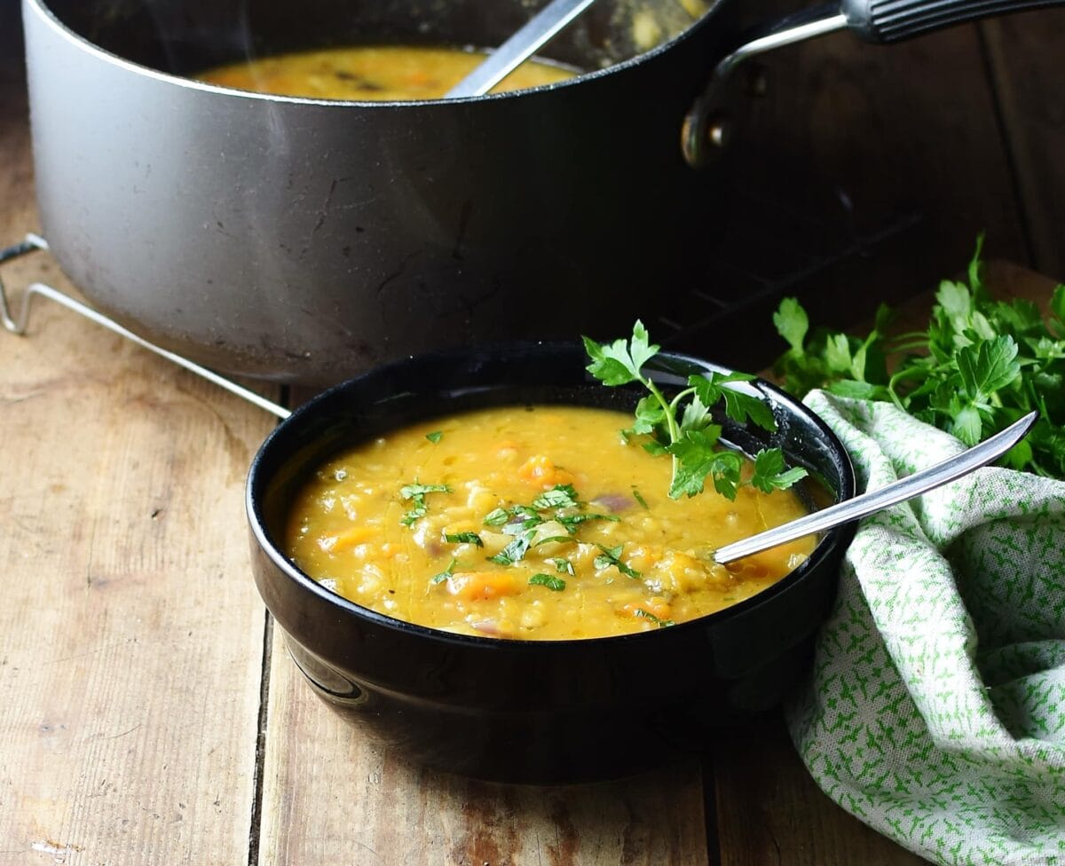 Side view of chunky soup with parsley and spoon in black bowl, green cloth and black pot with soup in background.
