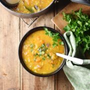 Chunky thick vegetable soup with chopped parsley and spoon in black bowl, with green cloth and parsley to the right and soup in pot at the top.