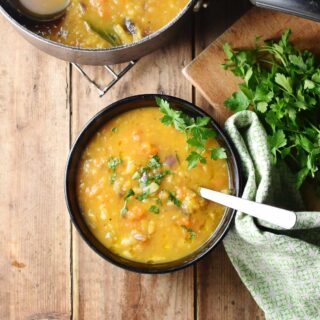 Chunky thick vegetable soup with chopped parsley and spoon in black bowl, with green cloth and parsley to the right and soup in pot at the top.