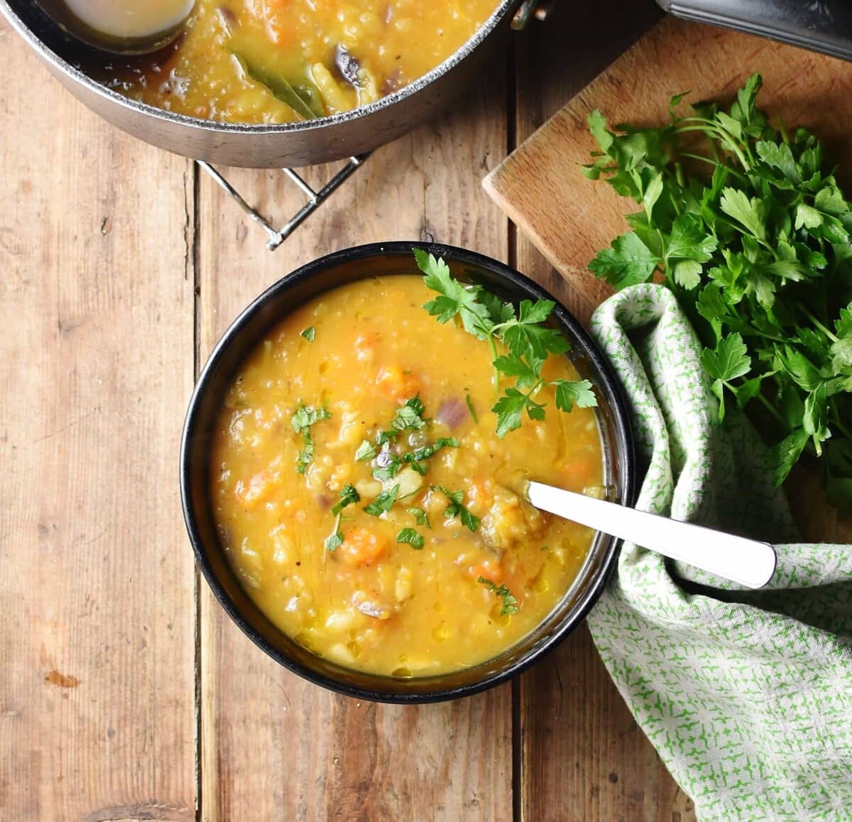 Chunky thick vegetable soup with chopped parsley and spoon in black bowl, with green cloth and parsley to the right and soup in pot at the top.