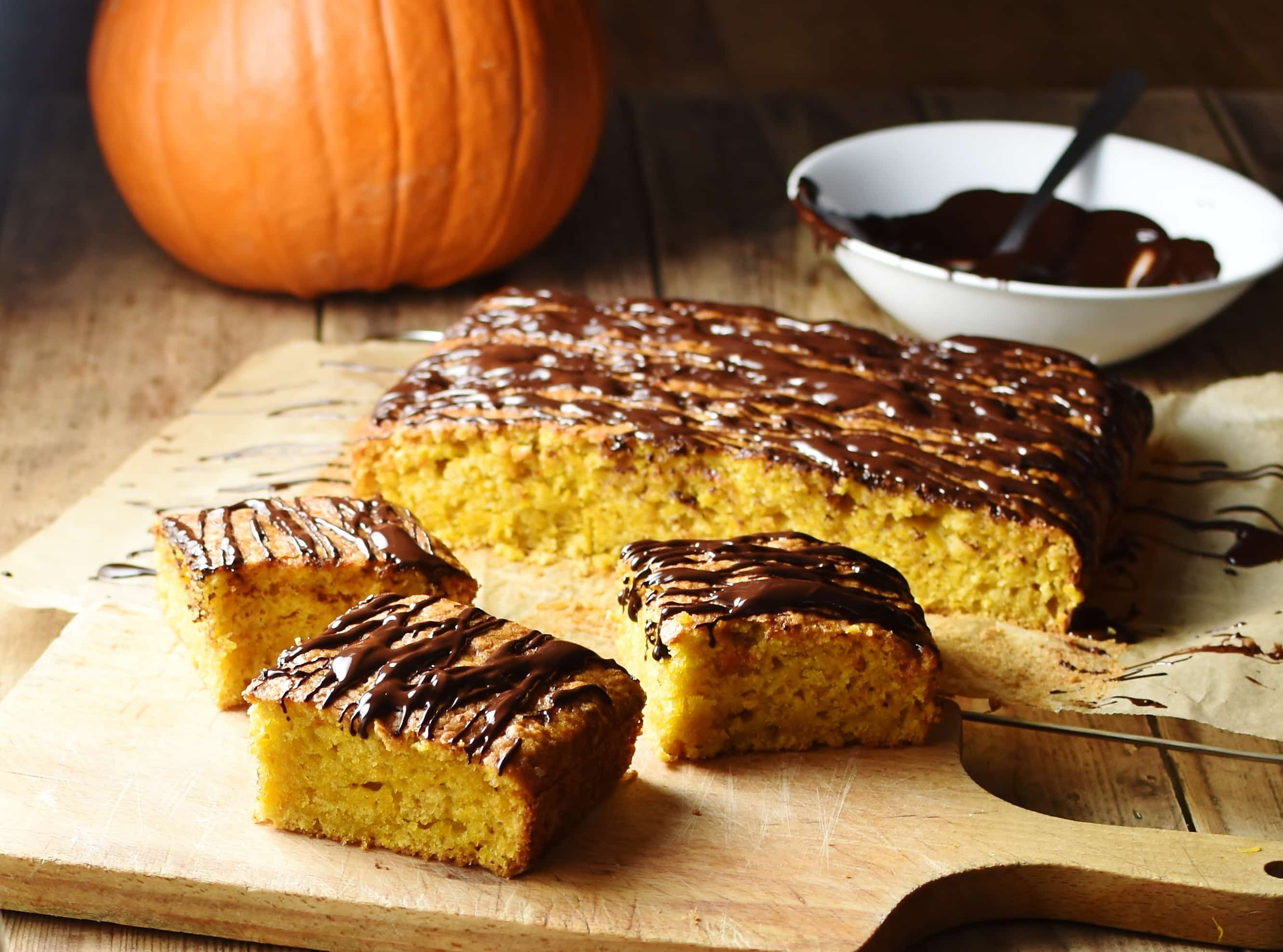 Pumpkin cake slices with chocolate drizzle and more cake in background, with pumpkin and melted chocolate in white bowl in top right.