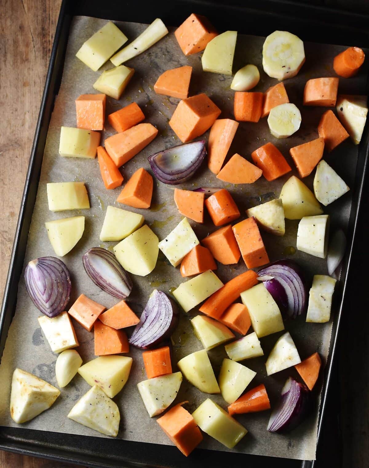 Chunks of peeled root vegetables and red onions on baking sheet lined with paper.
