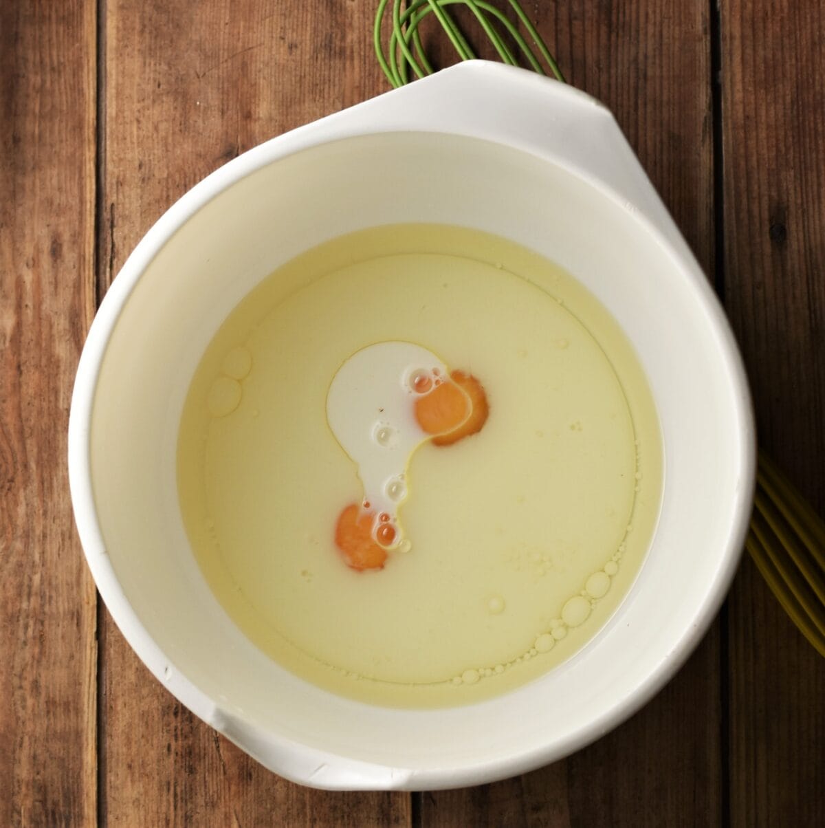 2 eggs and milk mixture in large white bowl with green whisk to the right.