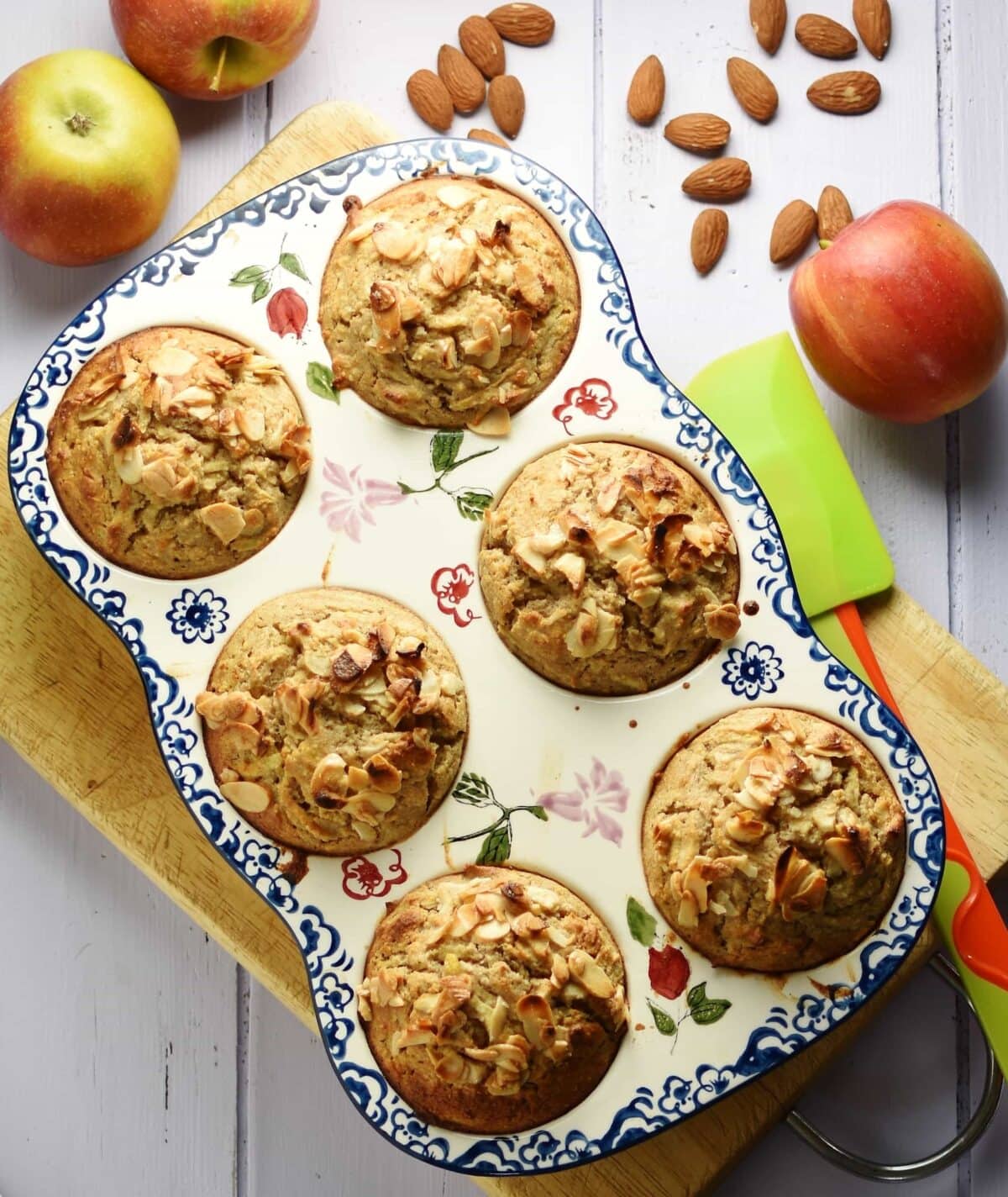 Top down view of muffins with almond flakes on top inside 6-hole ceramic muffin pan on top of cutting board, with green spatula, almonds and apples in background.