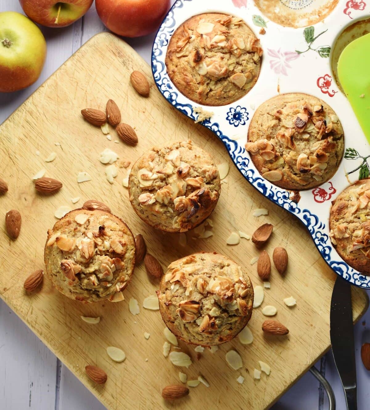Muffins with almond flakes and almonds on top of wooden board, and muffins in ceramic pan in background.