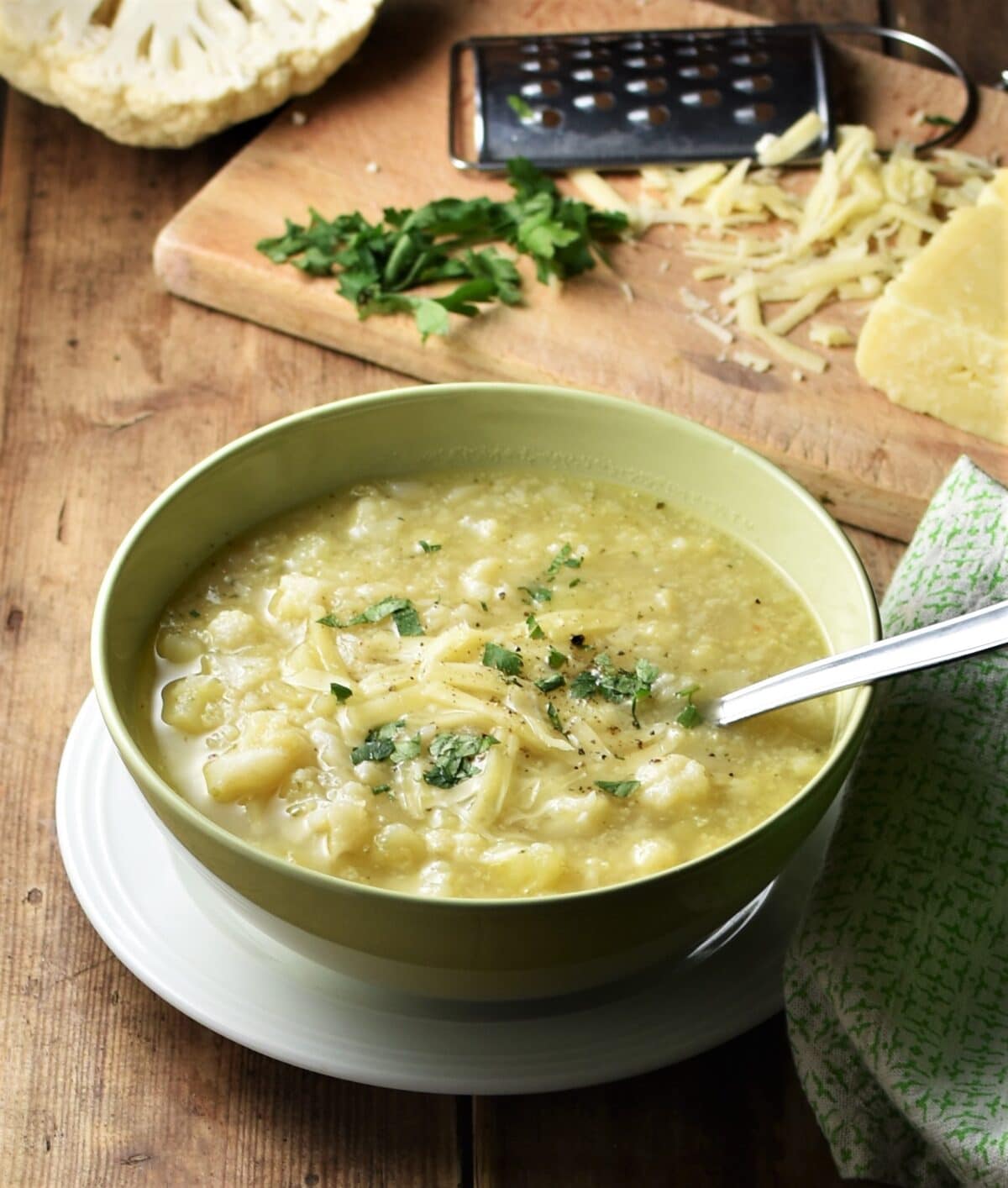 Chunky potato cauliflower soup in yellow bowl with spoon on white plate, with cauliflower, cheese, herbs and grater on top of wooden board and green cloth in background.