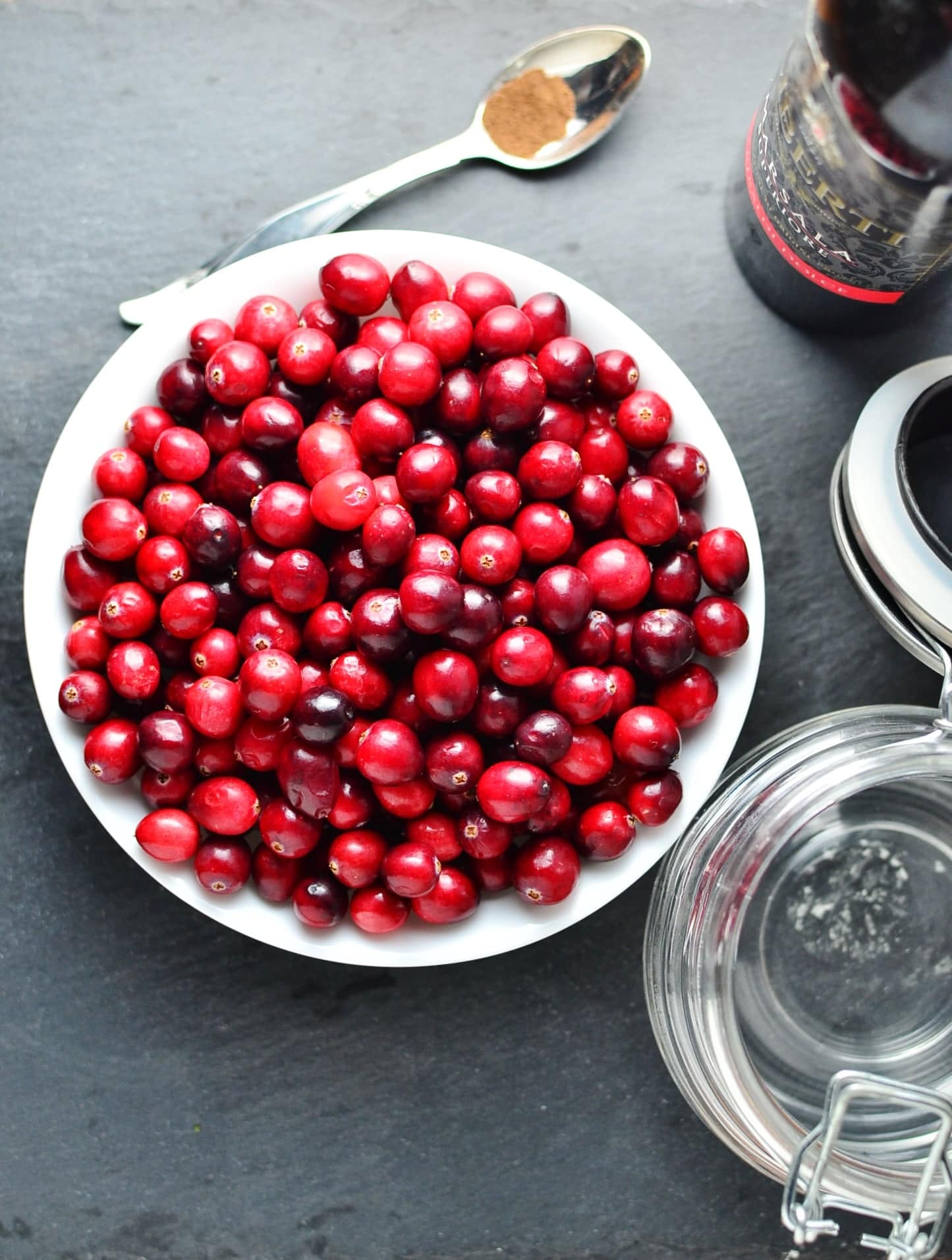 Top down view of raw cranberries inside white bowl, with spoon at top, dark bottle and open jar to the right.
