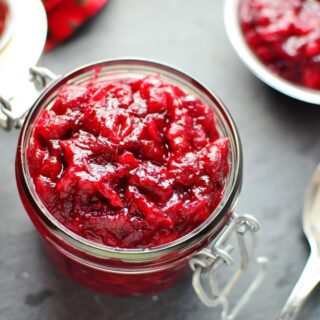 Top down view of cranberry sauce in open jar, with more cranberry sauce in background and spoon in bottom right.