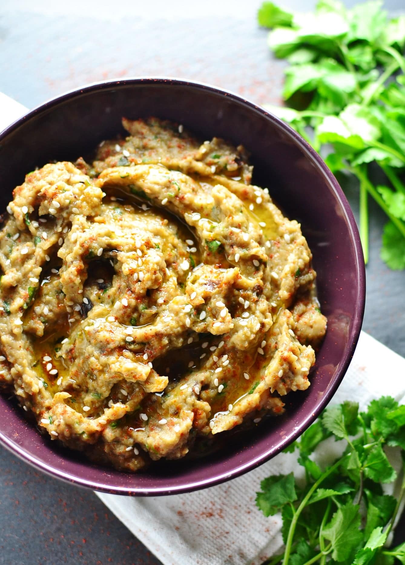 Roasted eggplant dip in purple bowl on white cloth with fresh cilantro on right.
