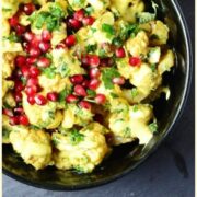 Top down view of curried cauliflower salad with yogurt dressing and pomegranate in black bowl, with chopped herbs and pomegranate seeds on top of wooden board