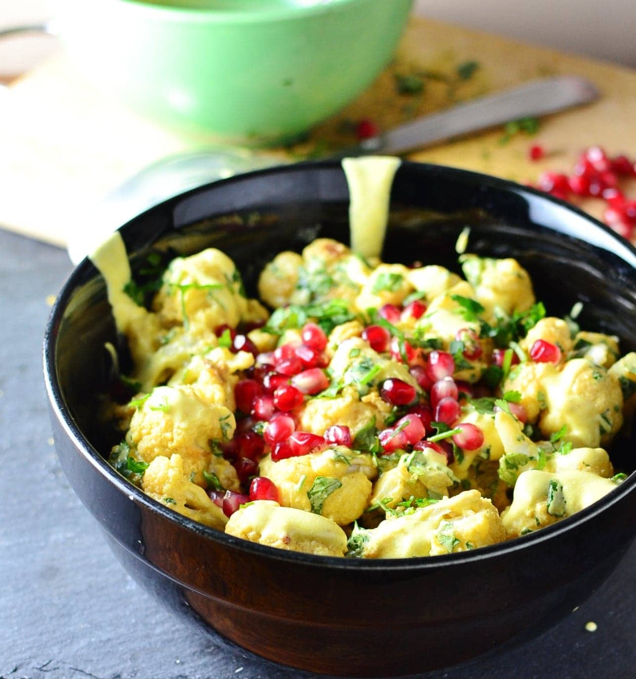 Curried cauliflower salad with yogurt dressing and pomegranate seeds in black bowl, with green bowl, chopped herbs and pomegranate seeds in background.