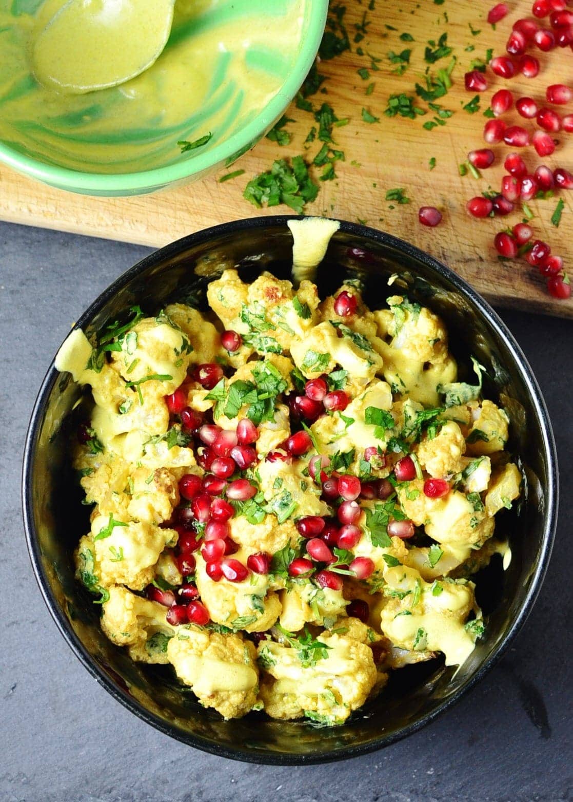 Top down view of curried cauliflower salad with yogurt dressing and pomegranate in black bowl, with green bowl, chopped herbs and pomegranate seeds on top of wooden board.