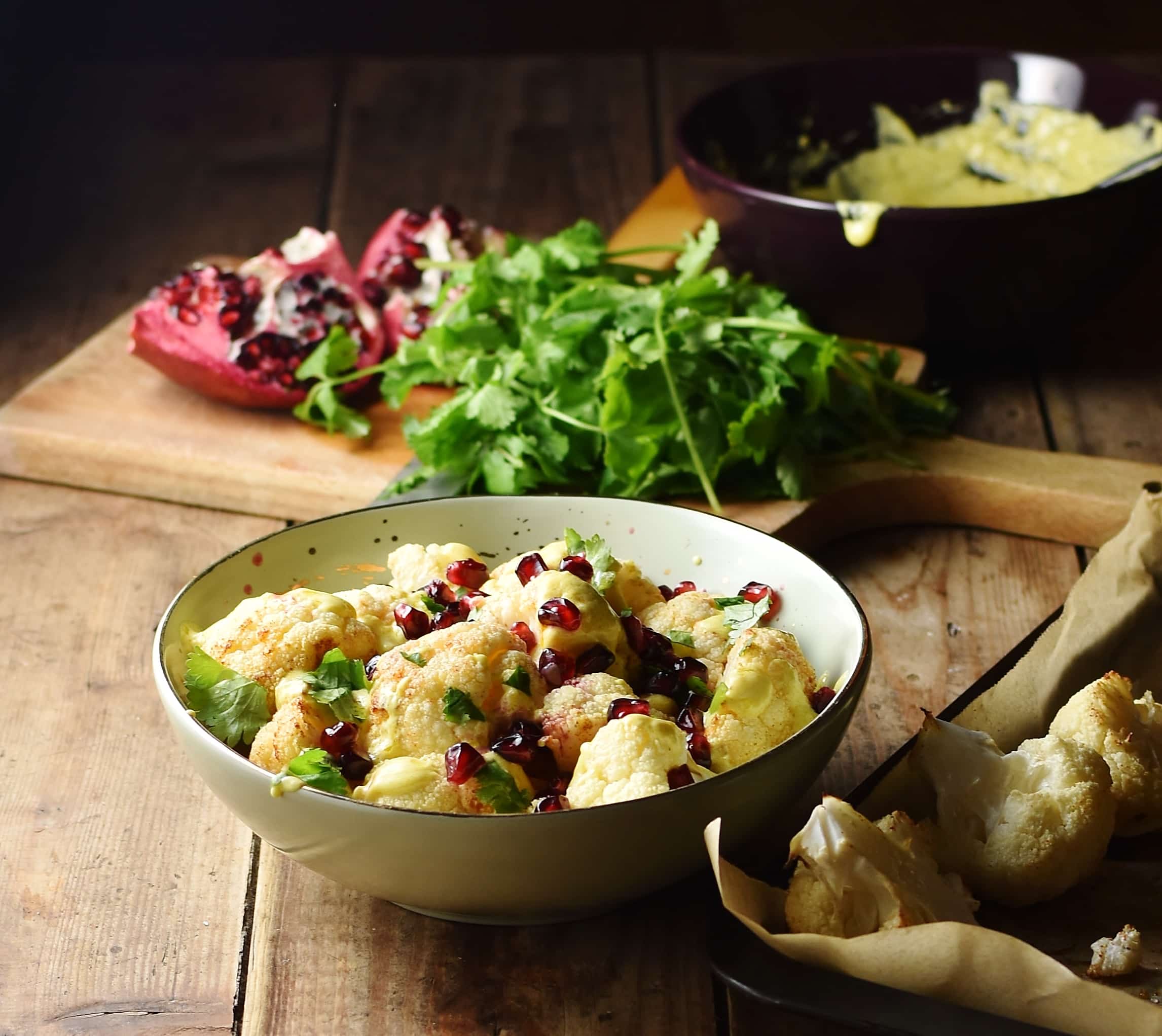 Side view of cauliflower salad with pomegranate seeds in yellow bowl with roasted cauliflower to the right, fresh herbs, pomegranate and creamy dressing in bowl in background.