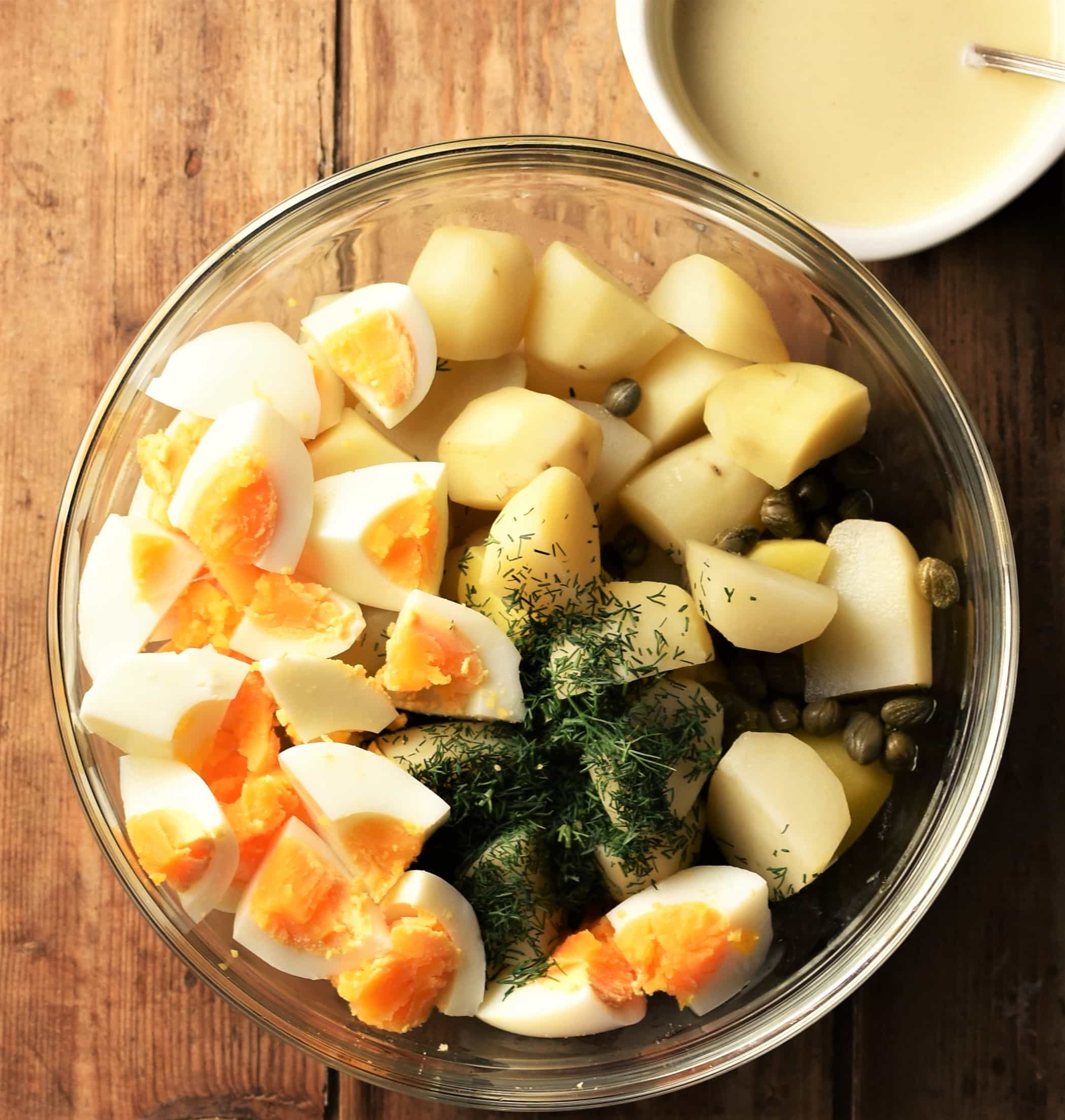 Cubed peeled potatoes and hard boiled egg, capers and dill in bowl with creamy dressing in top right.