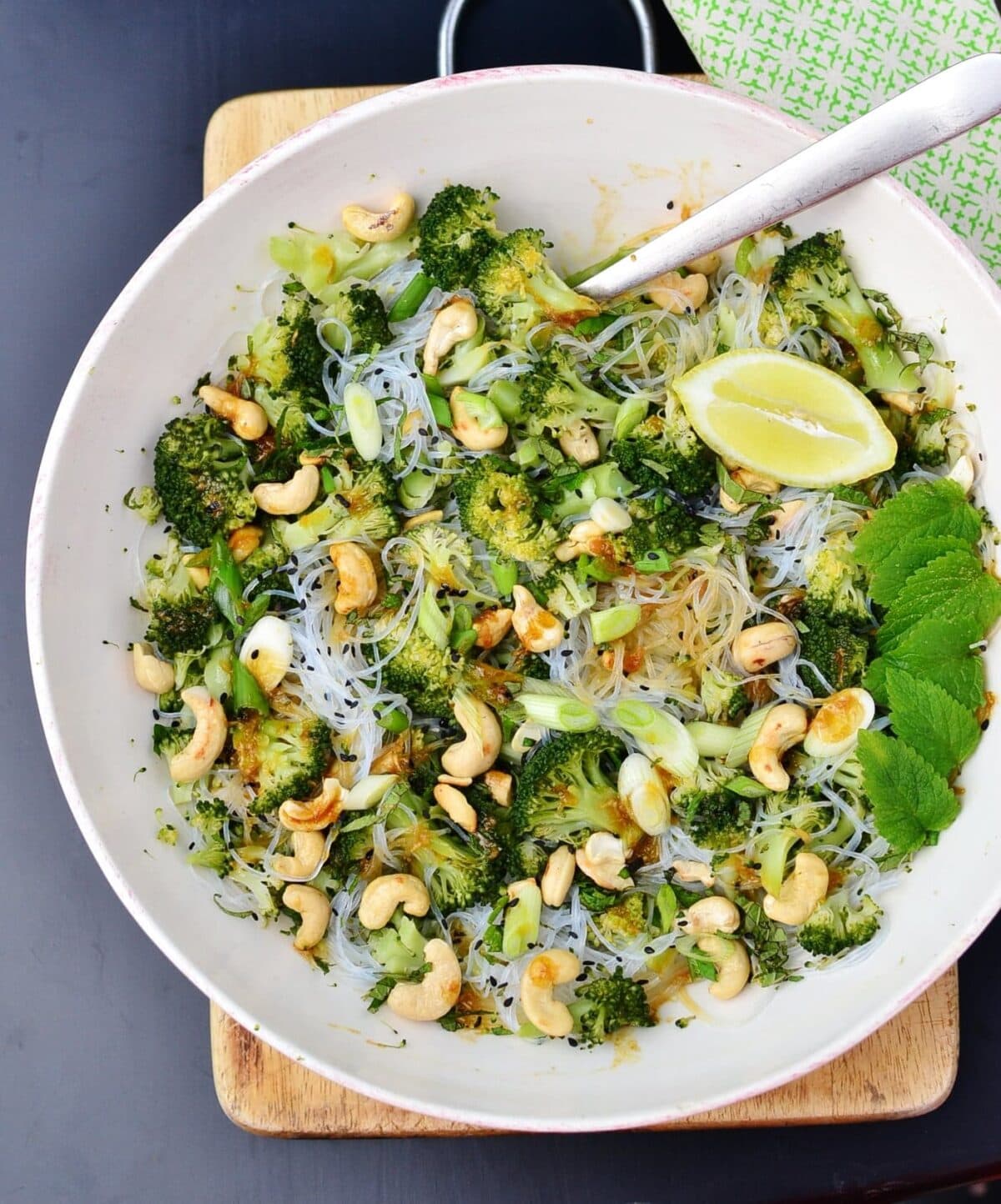 Top down view of bean thread noodles broccoli salad with cashews, lemon wedge, mint leaves and fork in white bowl, on top of wooden board, with green cloth to right.