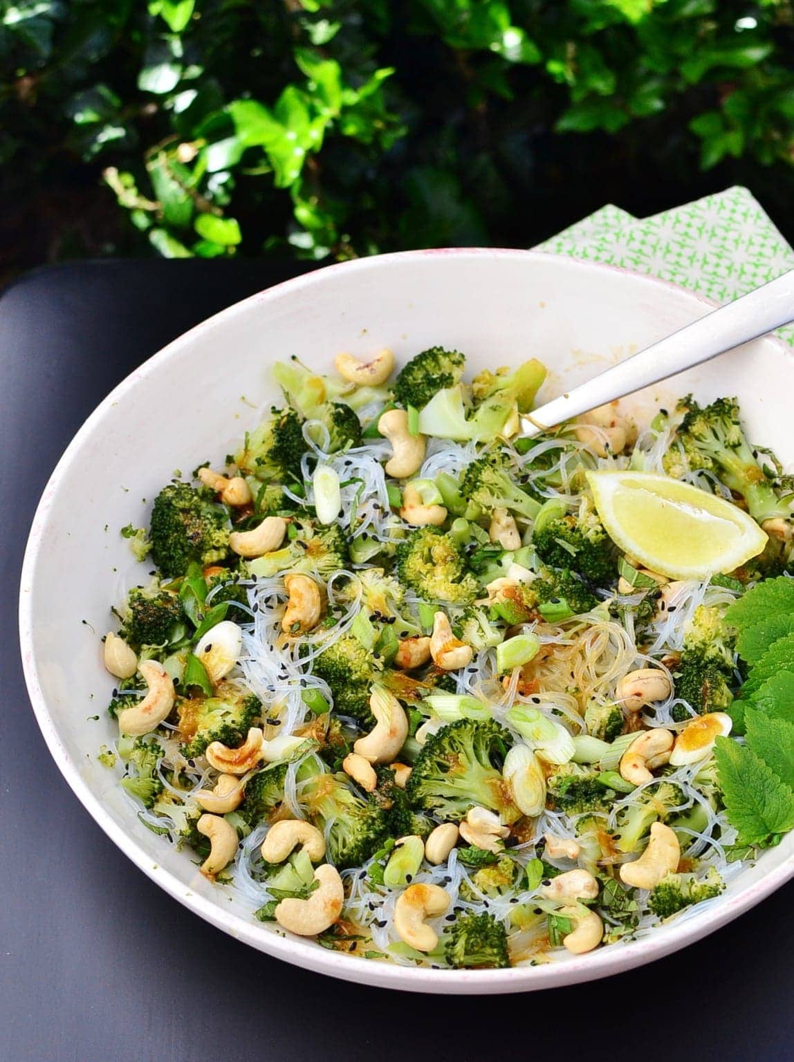 Close-up view of broccoli bean thread salad with cashews, mint leaves, lemon wedge and spoon inside white bowl.