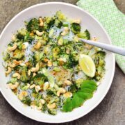 Top down view of bean thread noodles broccoli salad with cashews, lemon wedge, mint leaves and fork in white bowl with green cloth to right.