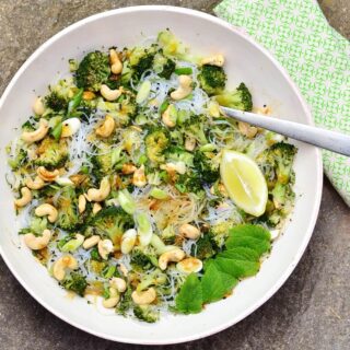 Top down view of bean thread noodles broccoli salad with cashews, lemon wedge, mint leaves and fork in white bowl with green cloth to right.