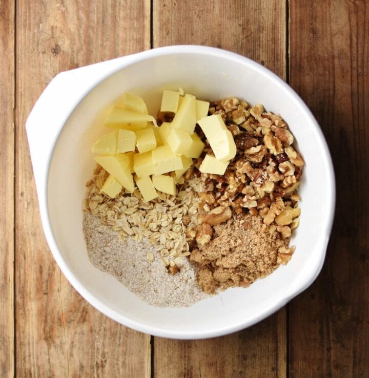 Cubed butter, oats, flour, walnuts and brown sugar inside large white bowl.