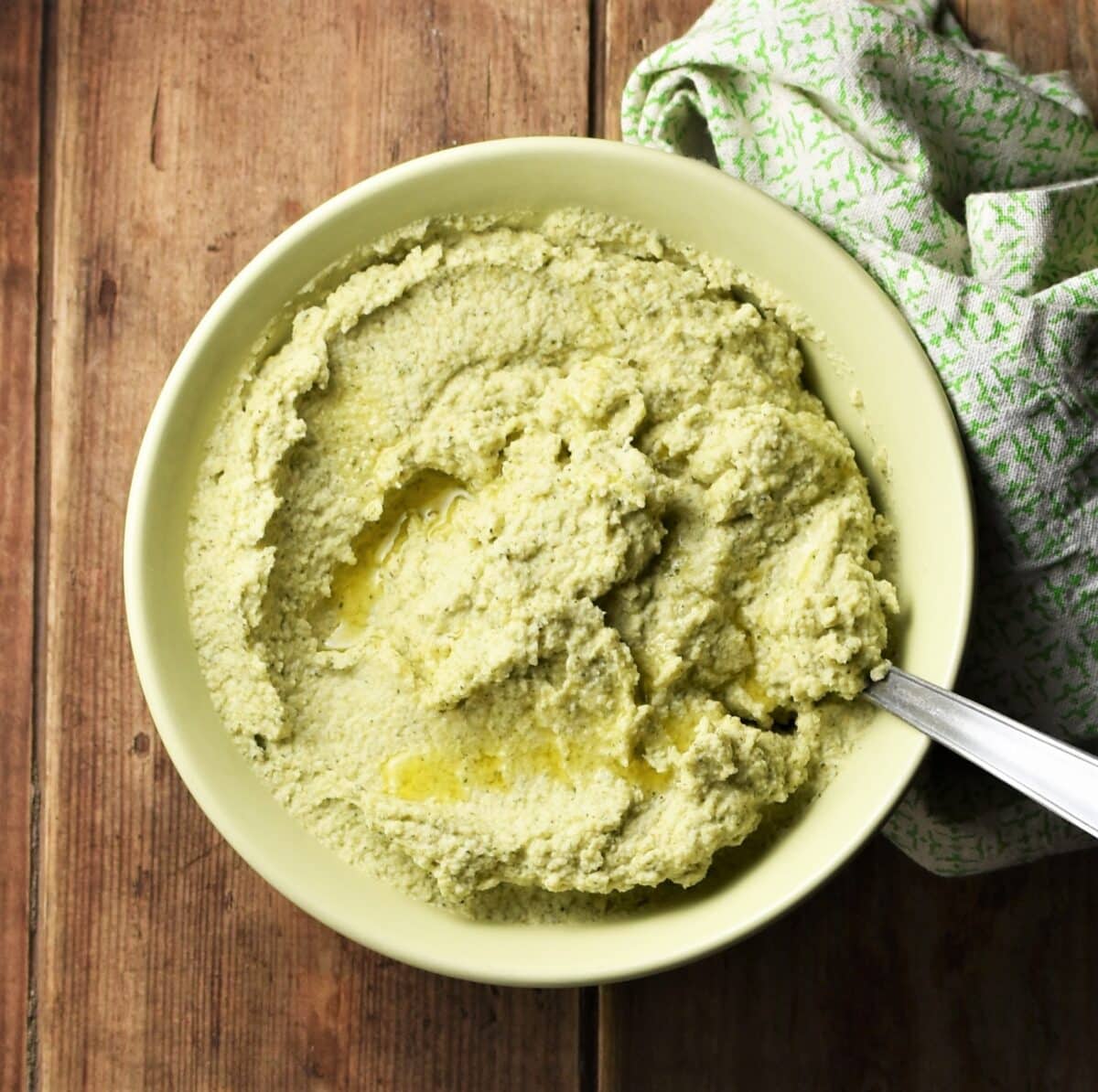 Top down view of broccoli dip with spoon in green bowl and green cloth to the right.