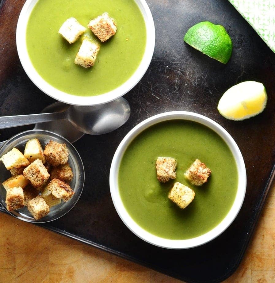 Top down view of split pea soup in 2 white bowls with croutons, lime wedges, spoons and dish of croutons on oven tray.