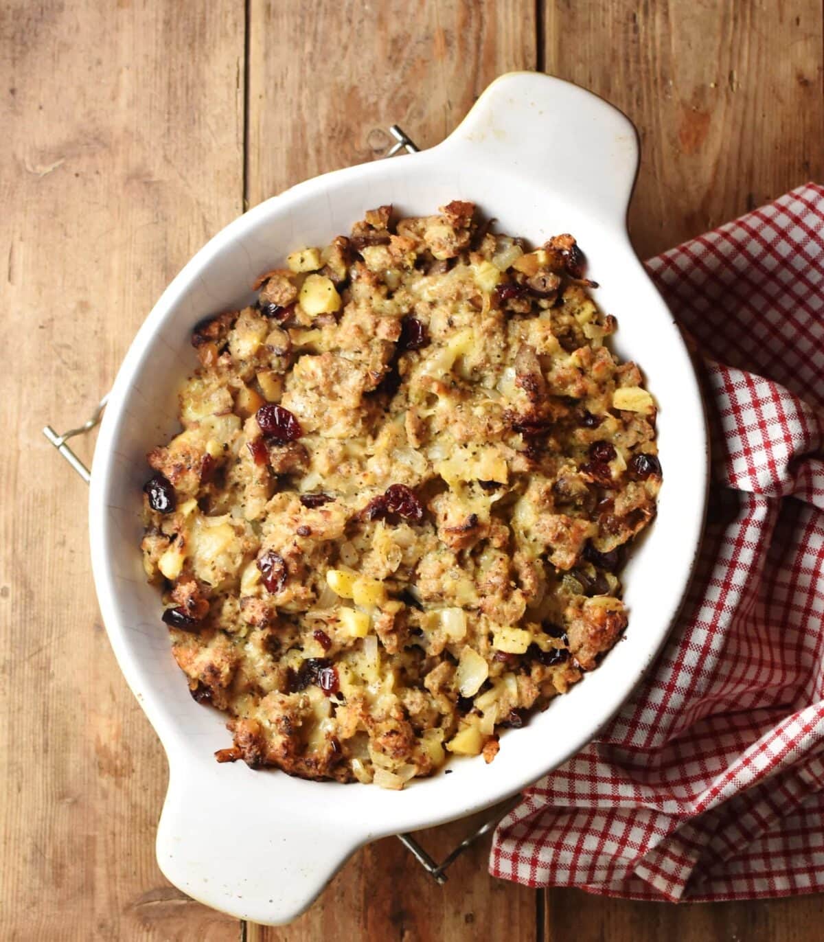 Top down view of baked vegetarian stuffing in white oval dish with red-and-white checkered cloth to the right.