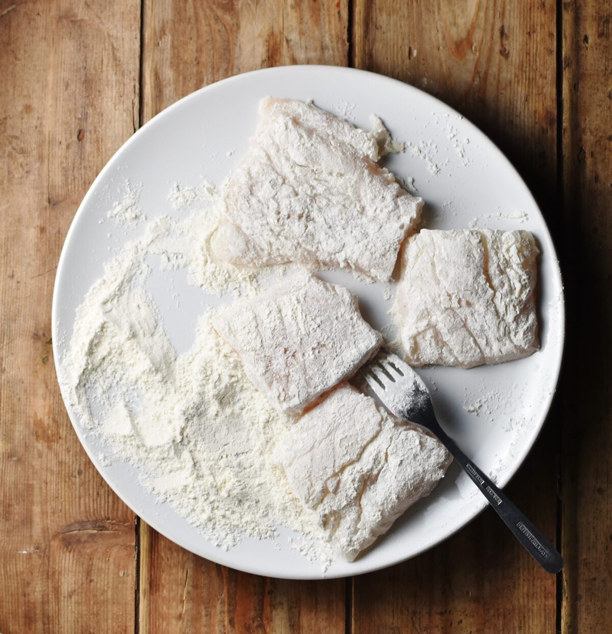 Fish fillets coated in flour on top of white plate with fork.