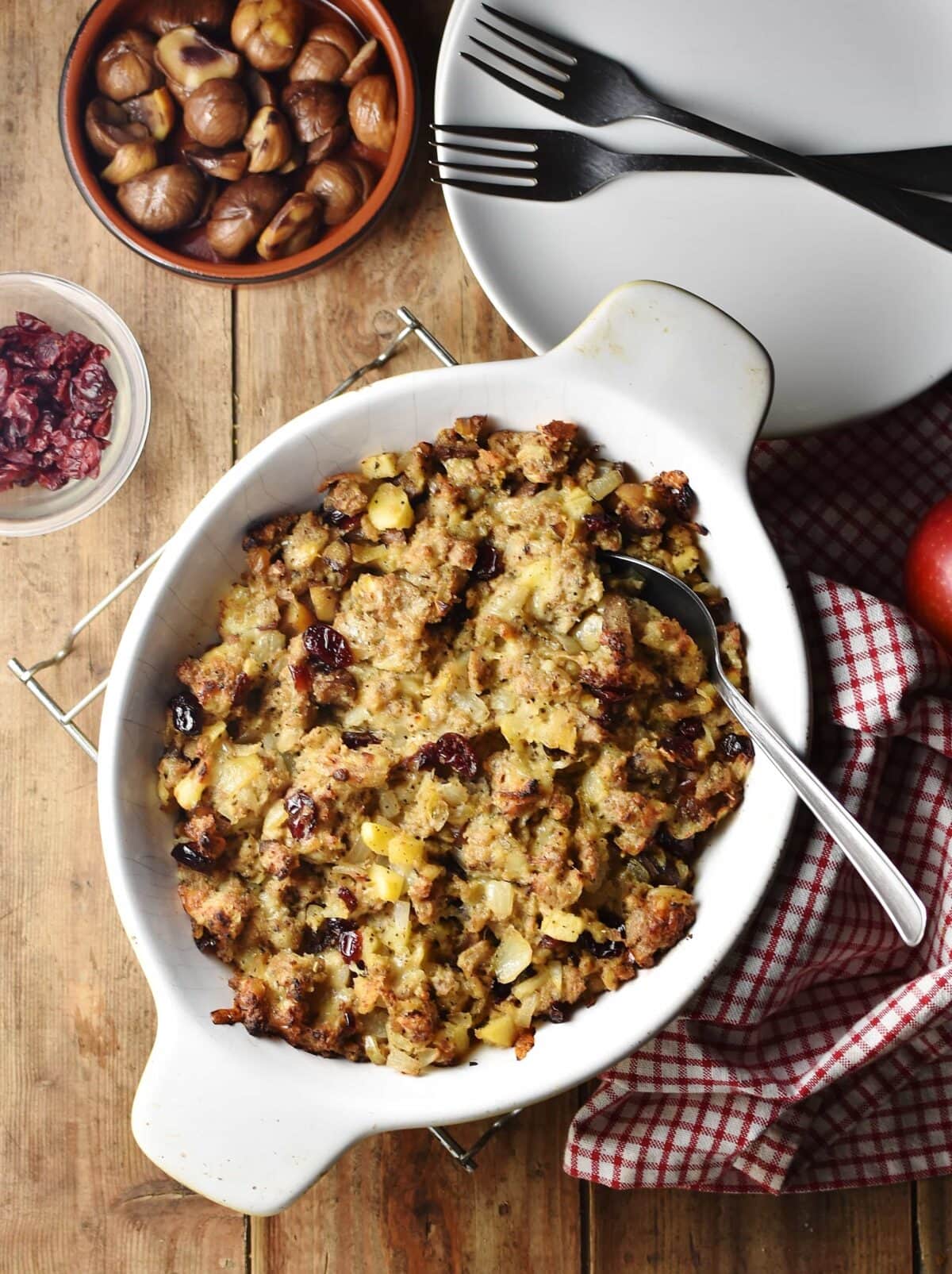 Christmas stuffing in white oval dish with spoon and red-and-white checkered cloth to the right, white plate with 2 forks in top right and chestnuts in round brown dish at the top.