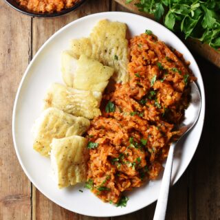 Top down view of fried fish fillets with stewed vegetables on white plate with large spoon, and herbs in background.