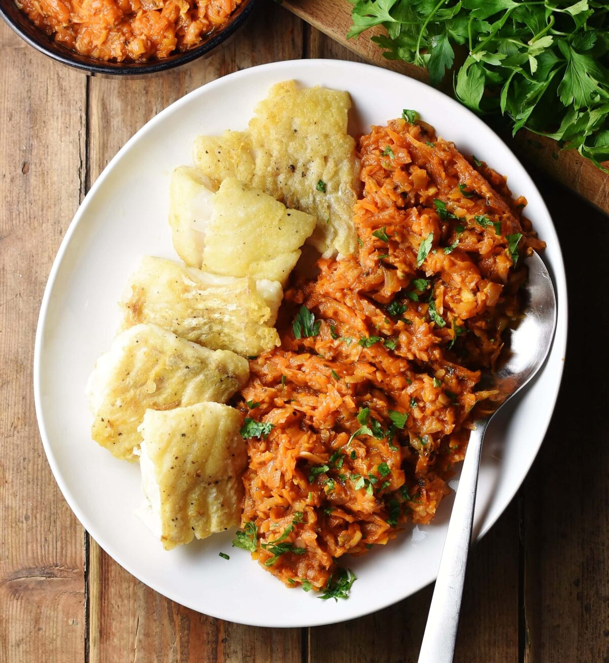 Top down view of fried fish fillets with stewed vegetables on white plate with large spoon, and herbs in background.