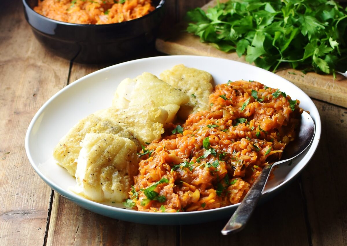 Stewed vegetables with fried fish on white plate with spoon, herbs and vegetable stew in black dish in background.