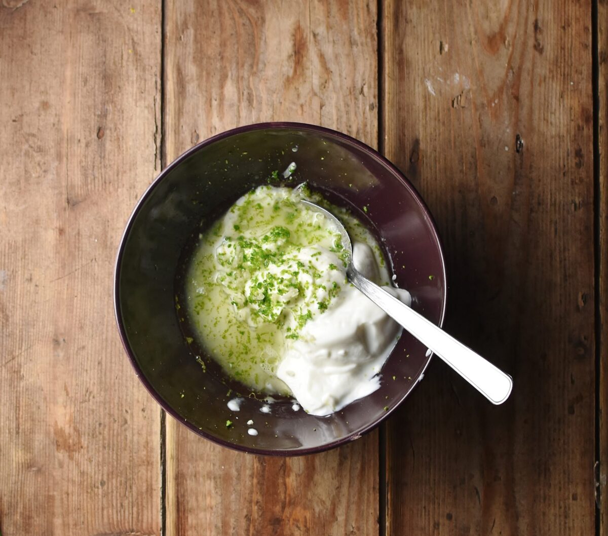 Top down view of yogurt, lime zest and oil in purple bowl with spoon. 