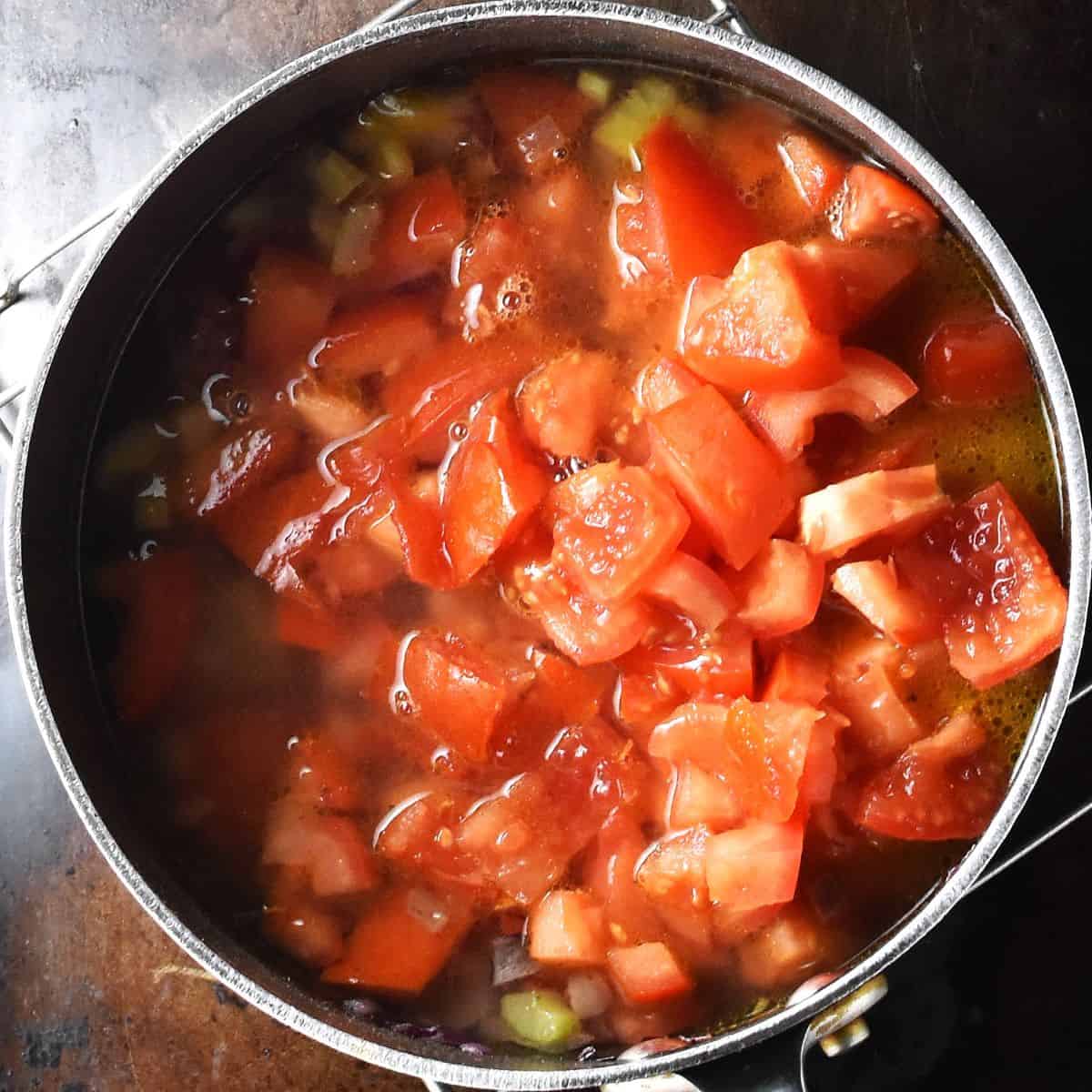 Adding chopped tomatoes and stock to soup in pot. 
