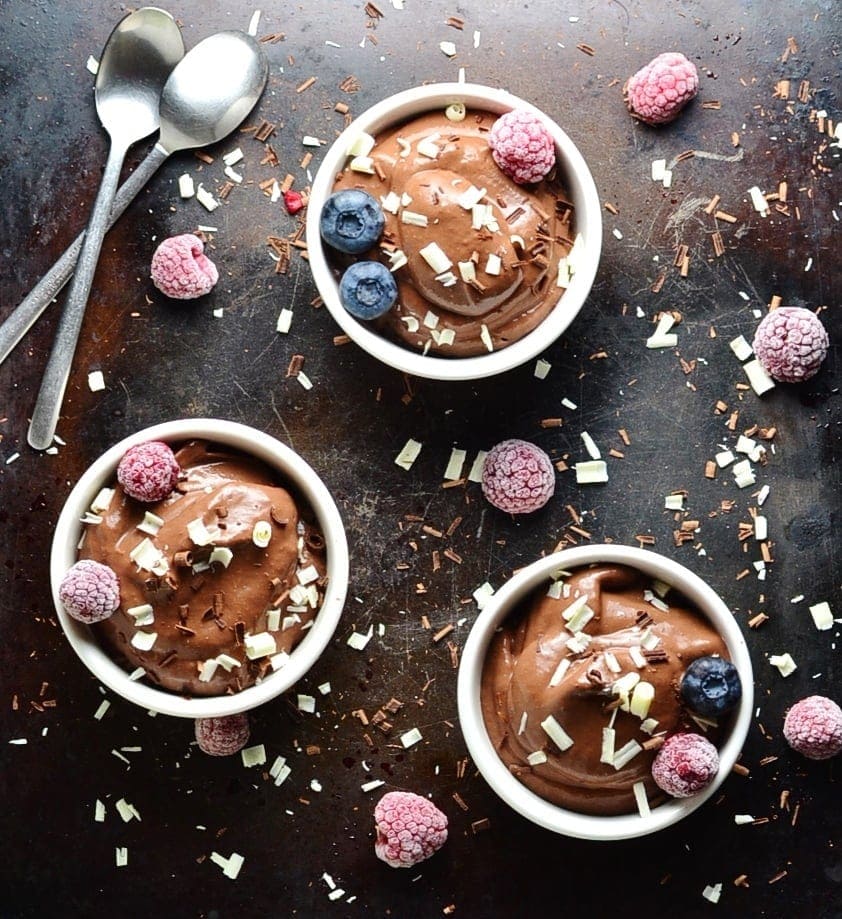 Top down view of chocolate banana mousse in white dishes with spoons, fruit and chocolate shavings on dark table.