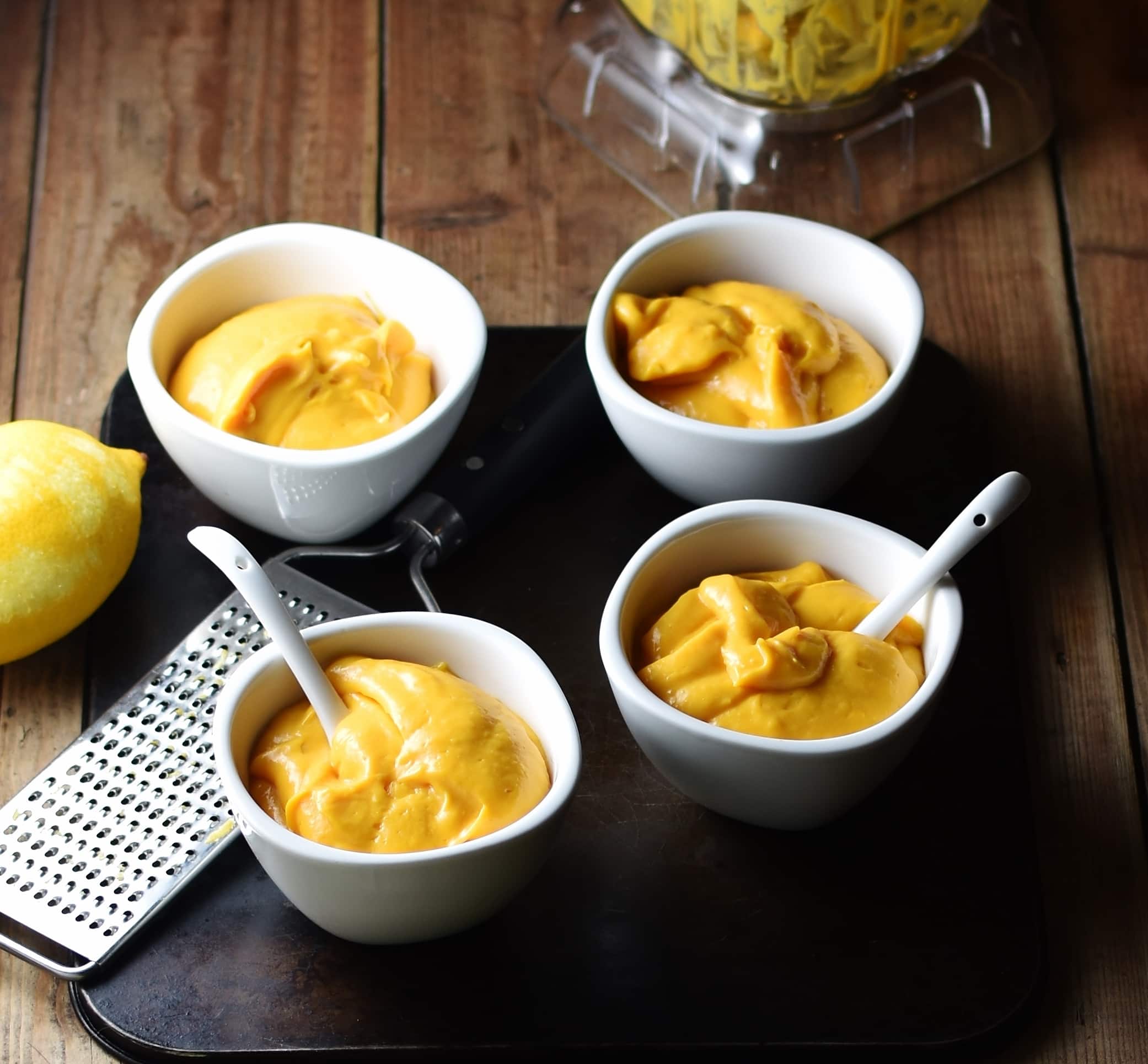 Sweet potato mousse in 4 white dishes with 2 white spoons, zester, lemon and blender in background.