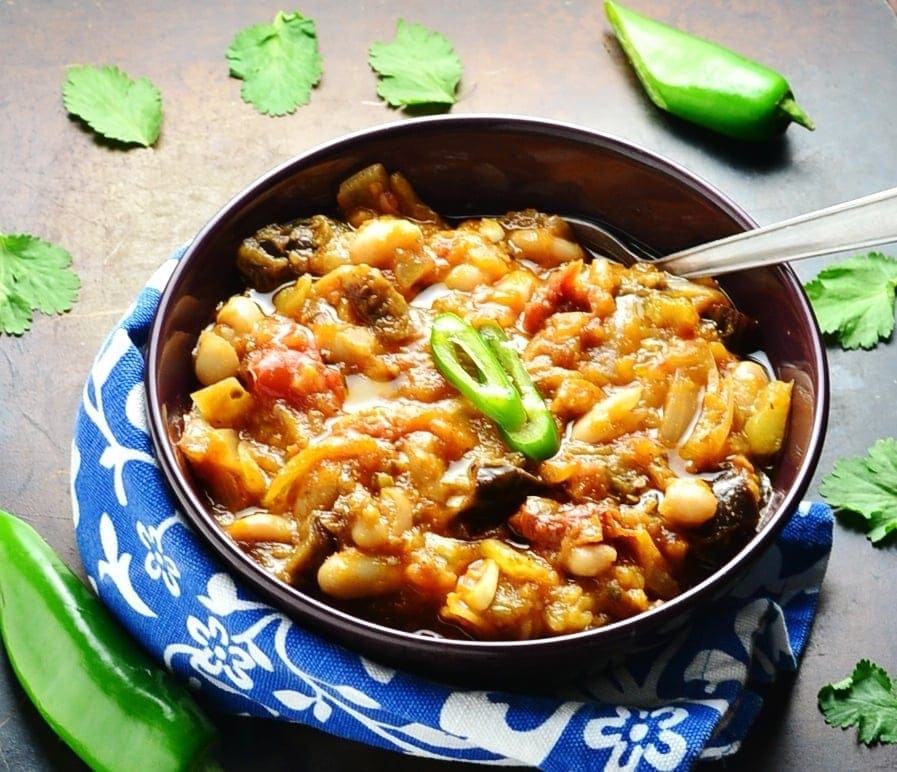 Eggplant casserole with spoon in purple bowl wrapped in blue-and-white cloth with green chillies and cilantro leaves on dark brown surface.
