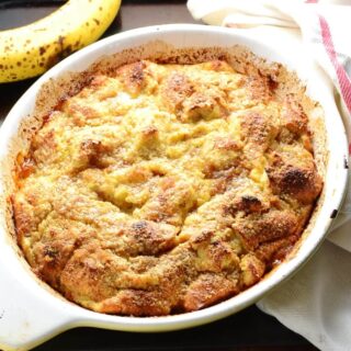 Banana french toast bake in white oval dish with ripe banana at top and white cloth with red stripe to right.