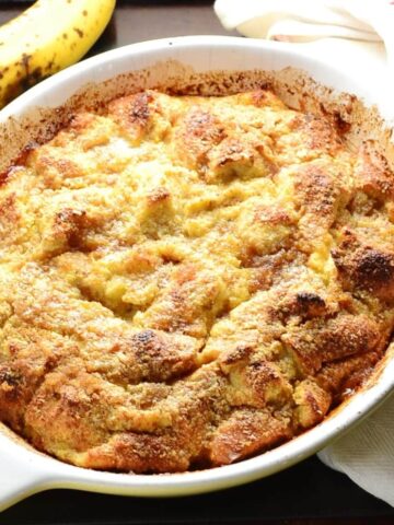 Banana french toast bake in white oval dish with ripe banana at top and white cloth with red stripe to right.