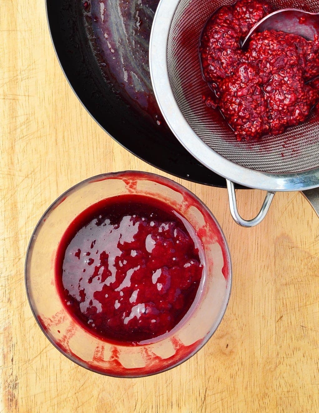 Top down view of raspberry sauce in small bowl and partial view of raspberry pulp inside sieve.