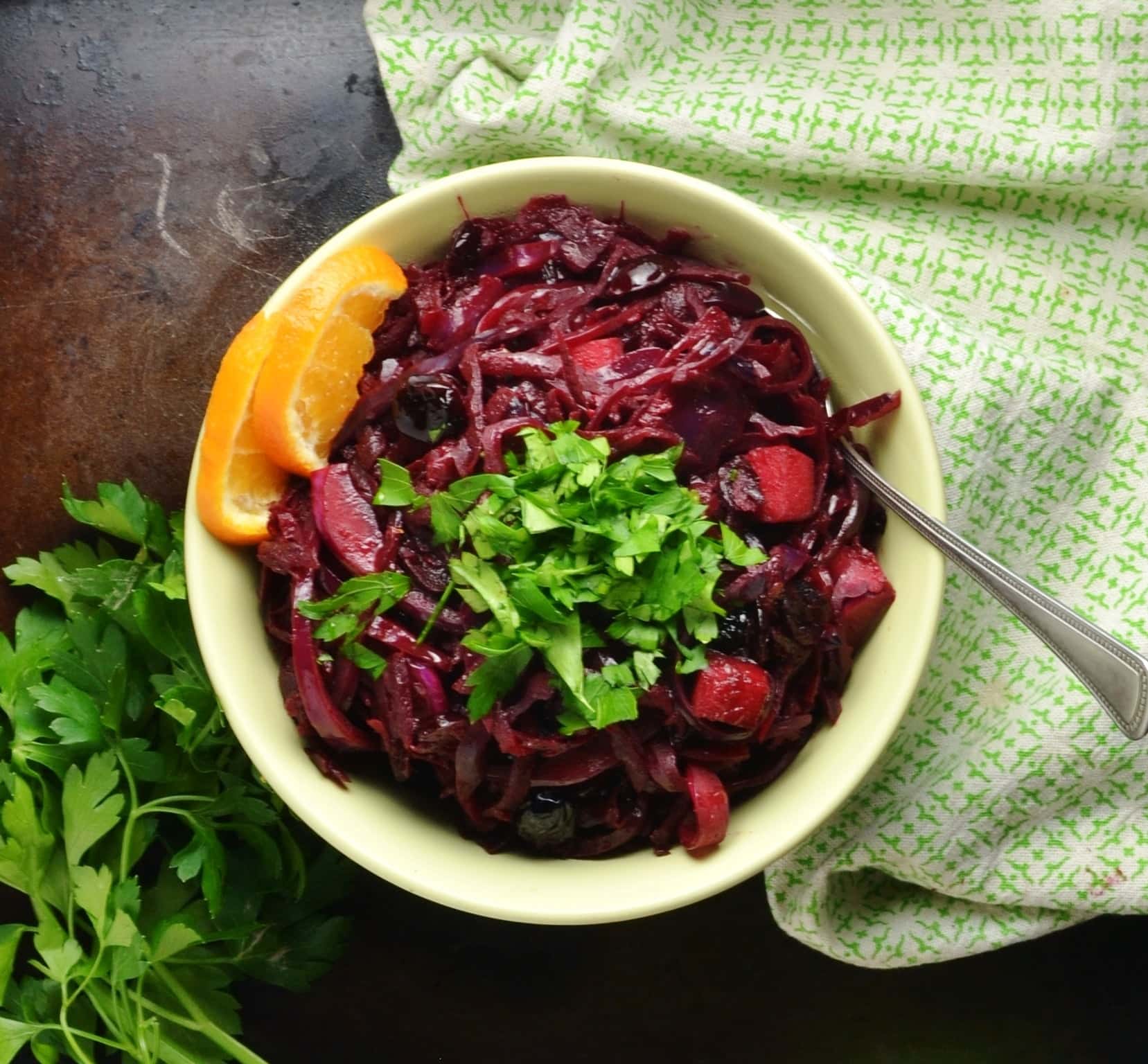 Top down view of chopped red cabbage in green bowl with spoon, herbs and green cloth.