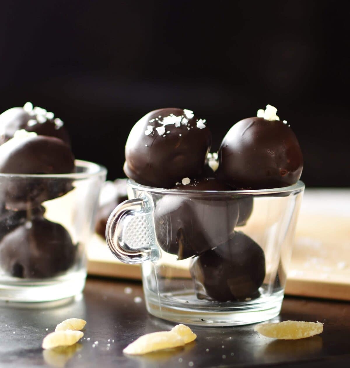Side view of chocolate truffles in 2 colourless cups with crystallised ginger in front and wooden board in background.