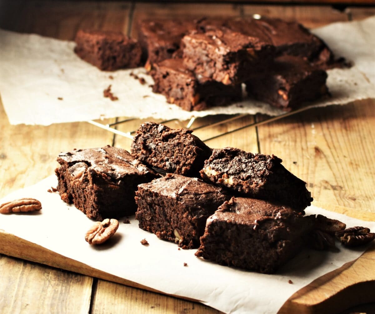 Brownies on top of paper with pecans and brownies in background on cooling rack.