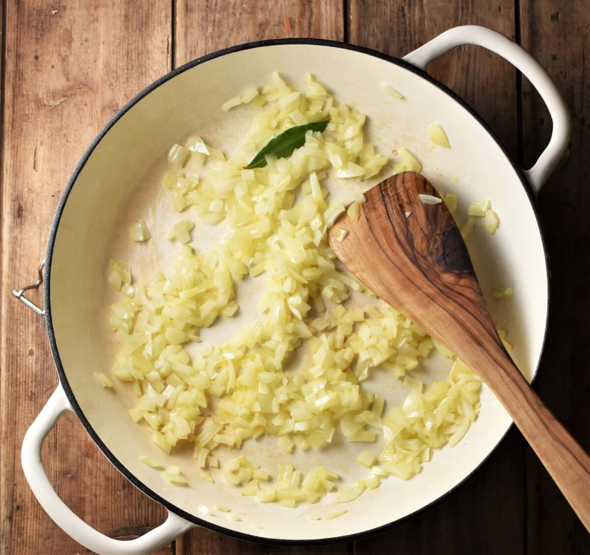 Chopped onion and bay leaf in large shallow white pan with wooden spoon.