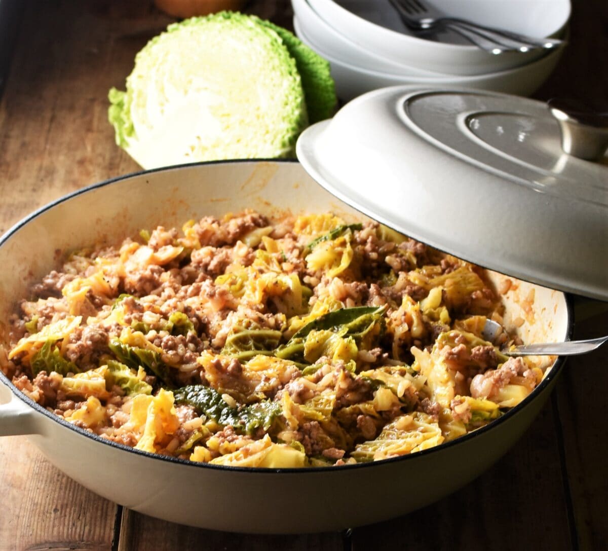 Side view of chopped cabbage, meat and rice in large white shallow dish with spoon and partly closed lid, with cabbage and white bowls in background.